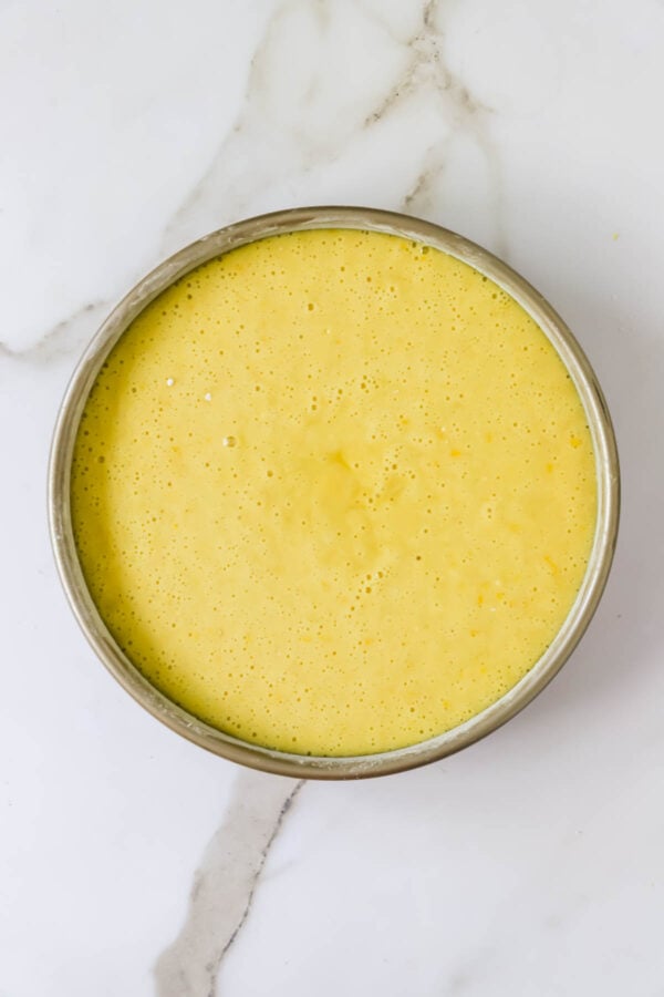 Round metal pan filled with a yellow, smooth, and bubbly liquid batter, placed on a white marble surface.