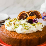 A round orange olive oil cake with whipped cream, chopped pistachios, and dried orange slices on top, displayed on a red plate.