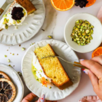 A hand cuts into a slice of orange olive oil cake with cream and pistachios on a white plate, surrounded by oranges, pistachios, and a pink napkin.
