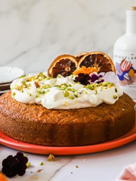 Round cake topped with whipped cream, chopped pistachios, edible flowers, and dried orange slices on a coral plate, with a bottle and small bowl nearby.