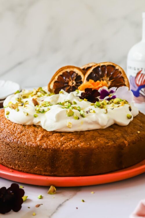 Round cake topped with whipped cream, chopped pistachios, edible flowers, and dried orange slices on a coral plate, with a bottle and small bowl nearby.