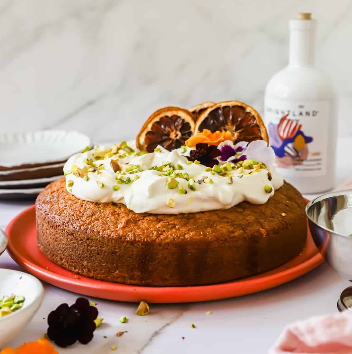 Round cake topped with whipped cream, chopped pistachios, edible flowers, and dried orange slices on a coral plate, with a bottle and small bowl nearby.