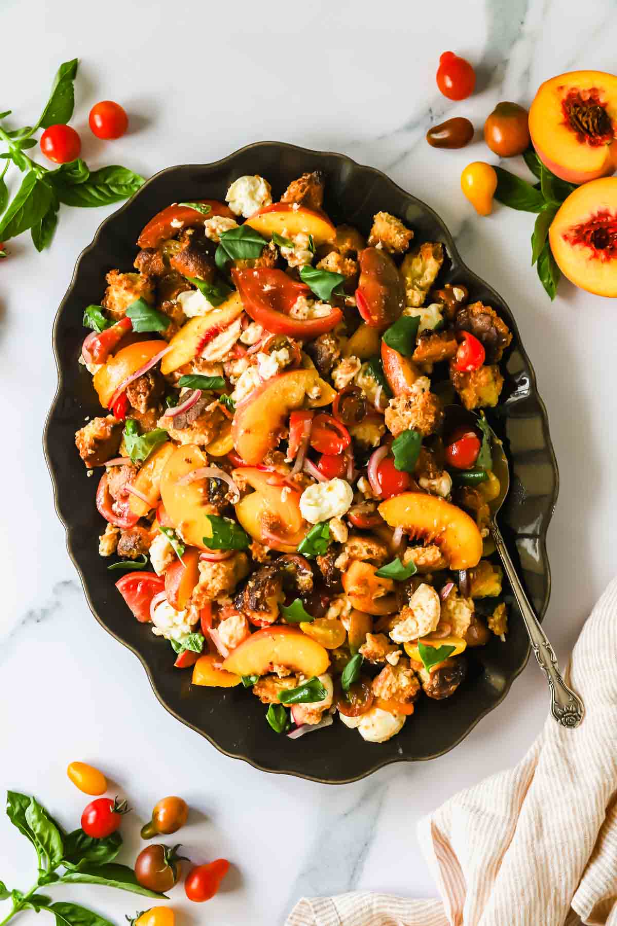 A black platter with panzanella salad featuring bread cubes, peaches, cherry tomatoes, mozzarella, fresh basil, and a few scattered ingredients on a marble surface.