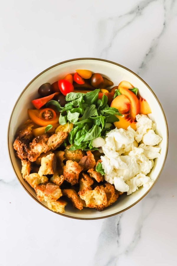 A bowl with torn bread, sliced cherry tomatoes, peach slices, mozzarella cheese, and fresh basil, arranged in separate sections.