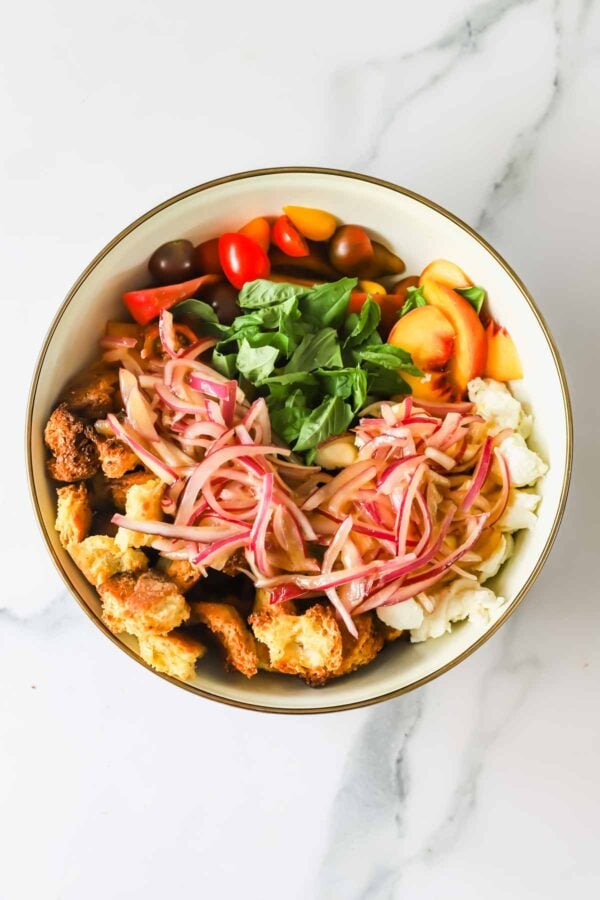 A bowl with cherry tomatoes, basil leaves, pickled red onions, chunks of bread, and pieces of mozzarella cheese on a marble surface.