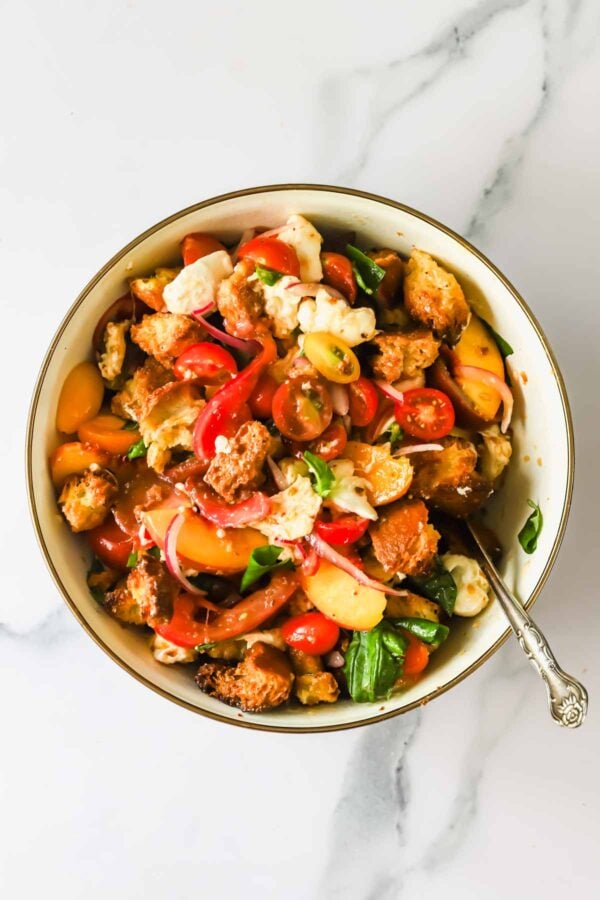 A bowl of panzanella salad with tomatoes, croutons, mozzarella, basil, and onions, on a marble surface with a fork in the bowl.