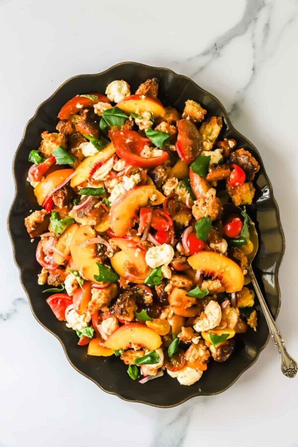 A black platter holds a salad of sliced peaches, tomatoes, mozzarella, bread cubes, basil, and a silver spoon, on a marble surface.