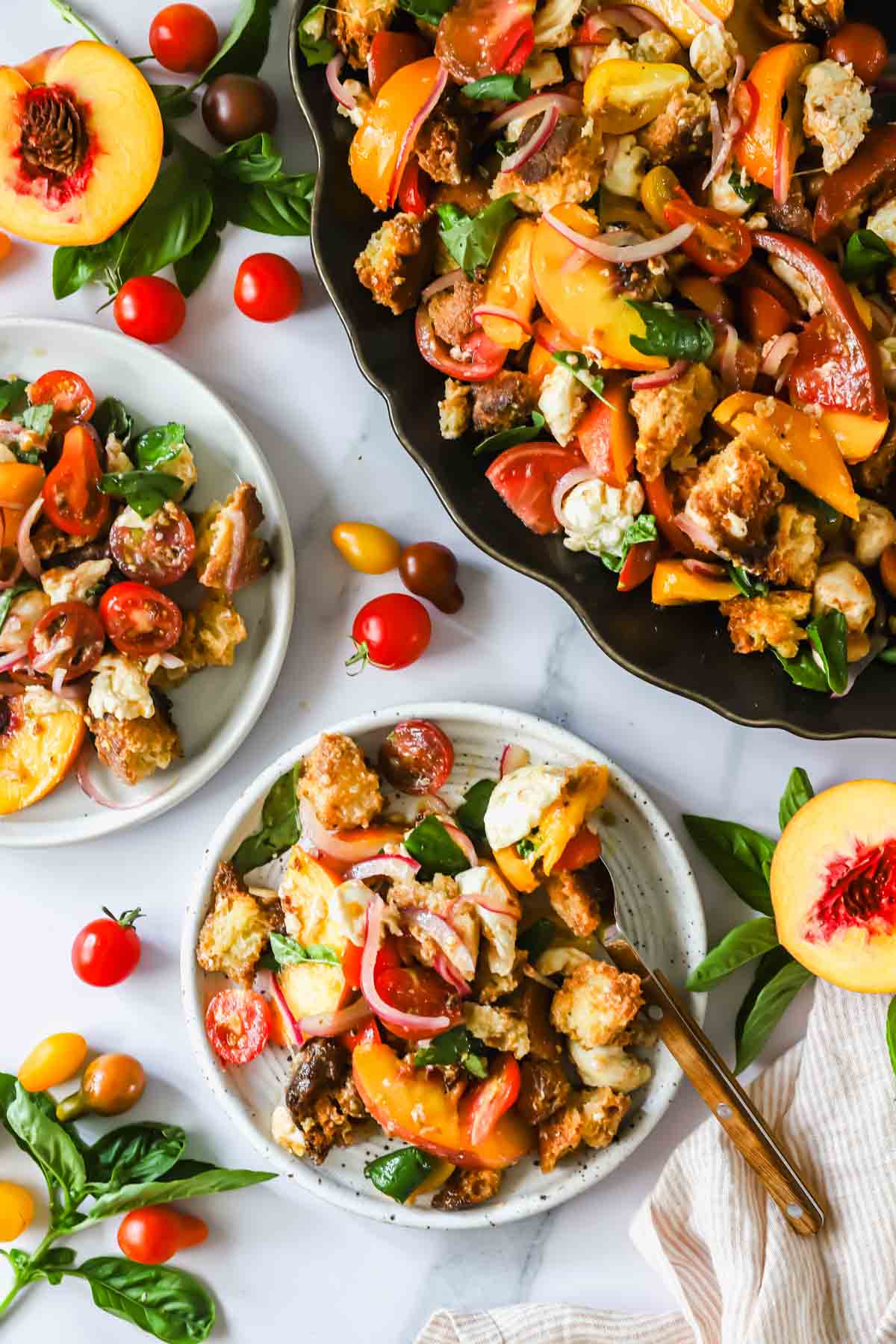 Plates and a platter of panzanella salad with tomatoes, peaches, basil, mozzarella, red onion, and bread pieces, surrounded by fresh ingredients on a white surface.