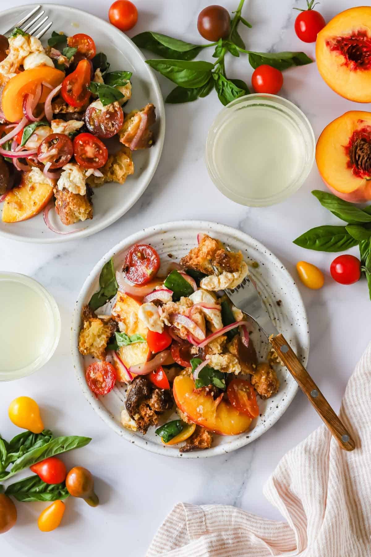 Two plates of salad with tomatoes, peaches, bread, basil, and cheese surrounded by fresh ingredients on a white surface, next to two glasses of a light beverage.