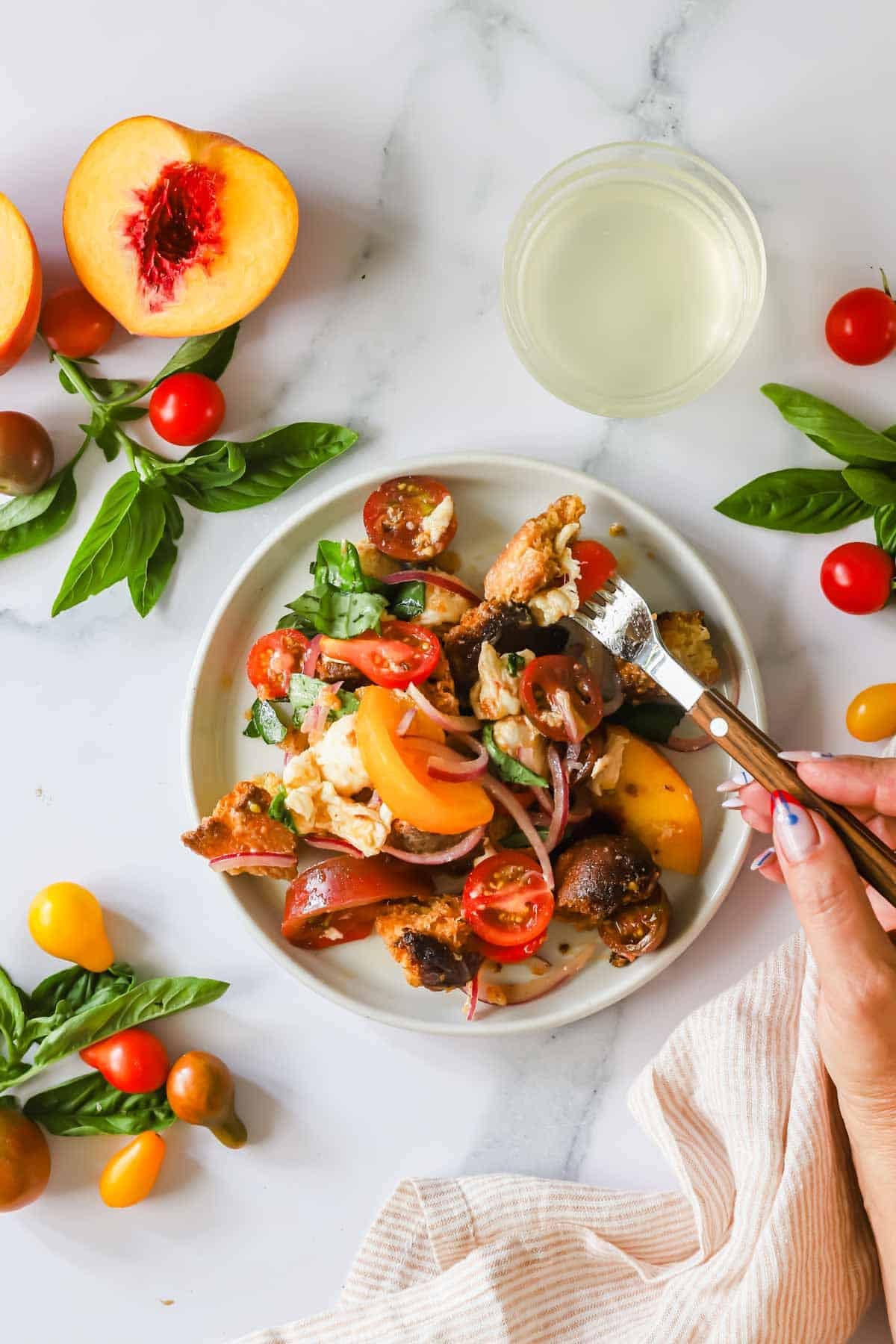 A plate of salad with tomatoes, peaches, red onions, basil, and bread pieces, next to a glass of white beverage and scattered fresh ingredients on a white marble surface.