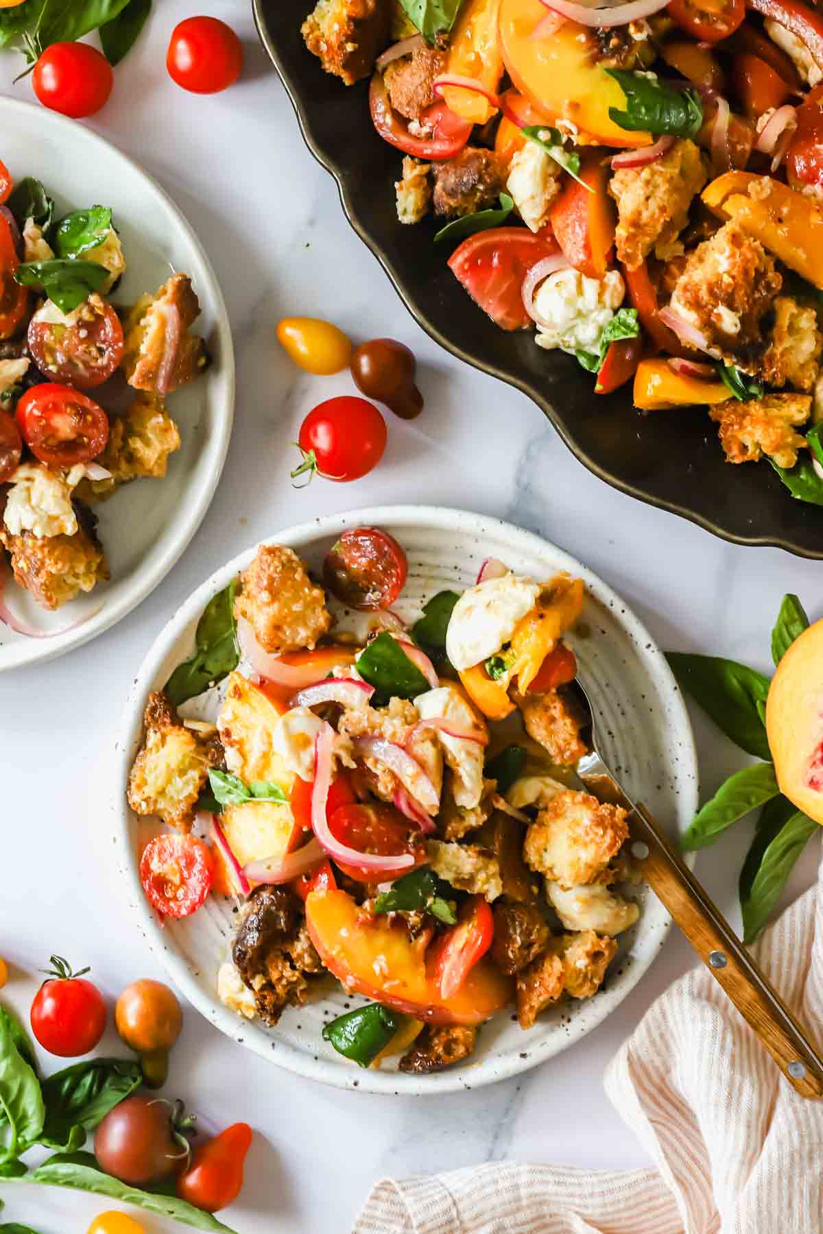 Plates of panzanella salad with tomatoes, bread, onions, and basil on a white surface, surrounded by scattered cherry tomatoes and basil leaves.