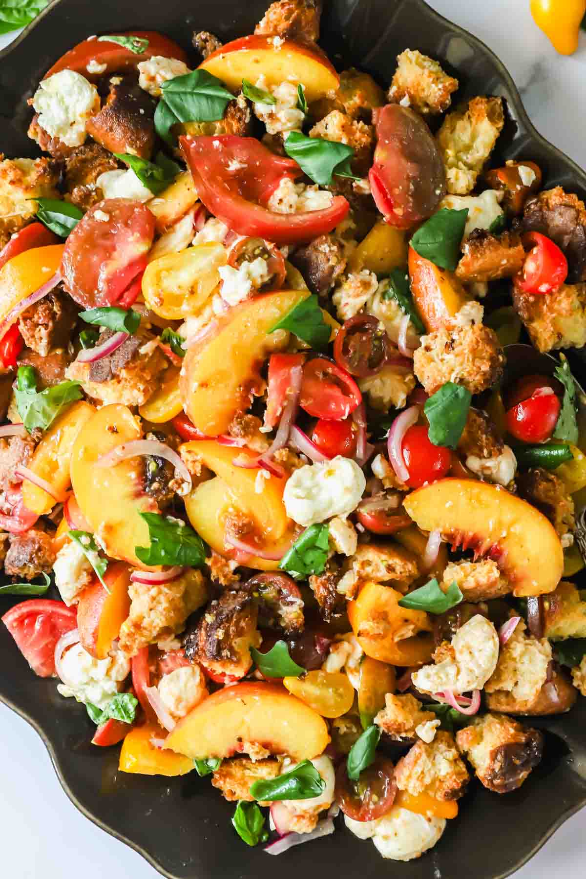 Salad with sliced tomatoes, peaches, chunks of bread, cheese, red onions, and fresh basil on a dark platter.