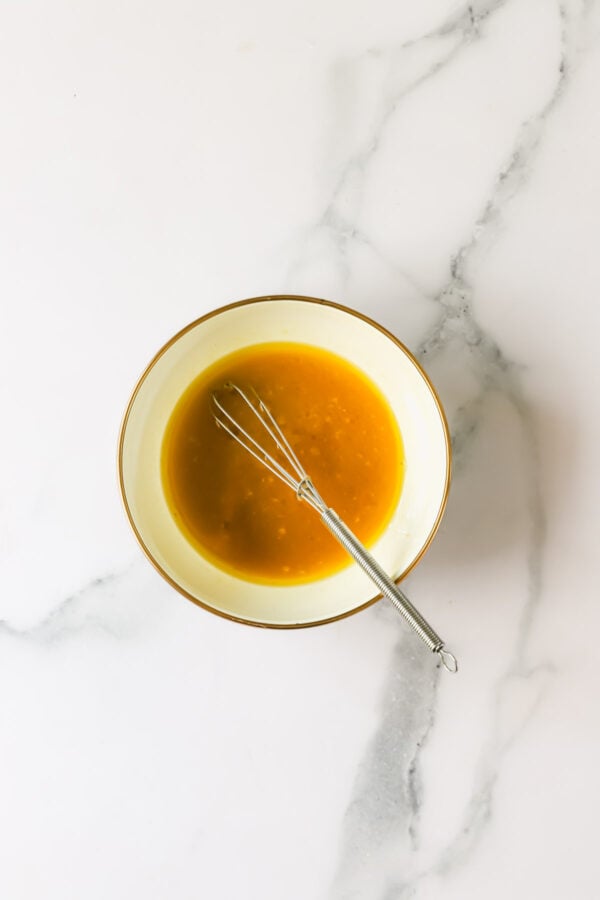 A small metal whisk rests inside a bowl filled with orange liquid, placed on a white marble surface.