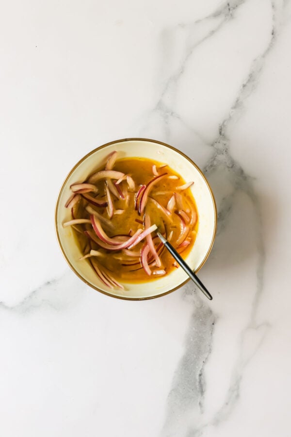 A bowl with sliced red onions in a brownish liquid, placed on a white marble surface with a spoon inside the bowl.