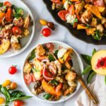 Three plates of panzanella salad with tomatoes, peaches, basil, croutons, and red onions are displayed on a white surface with basil leaves and cherry tomatoes scattered around.