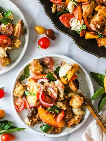 Three plates of panzanella salad with tomatoes, peaches, basil, croutons, and red onions are displayed on a white surface with basil leaves and cherry tomatoes scattered around.