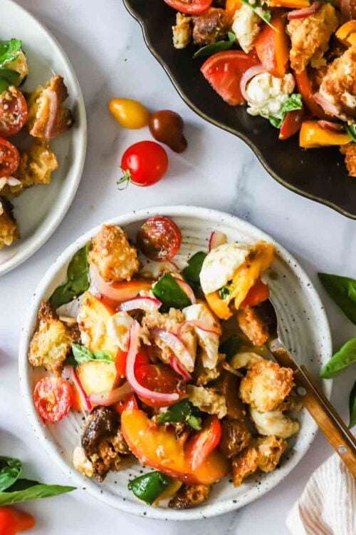 Three plates of panzanella salad with tomatoes, peaches, basil, croutons, and red onions are displayed on a white surface with basil leaves and cherry tomatoes scattered around.