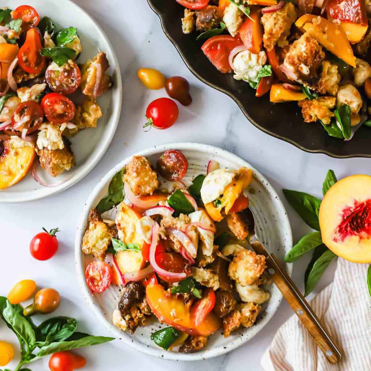 Three plates of panzanella salad with tomatoes, peaches, basil, croutons, and red onions are displayed on a white surface with basil leaves and cherry tomatoes scattered around.