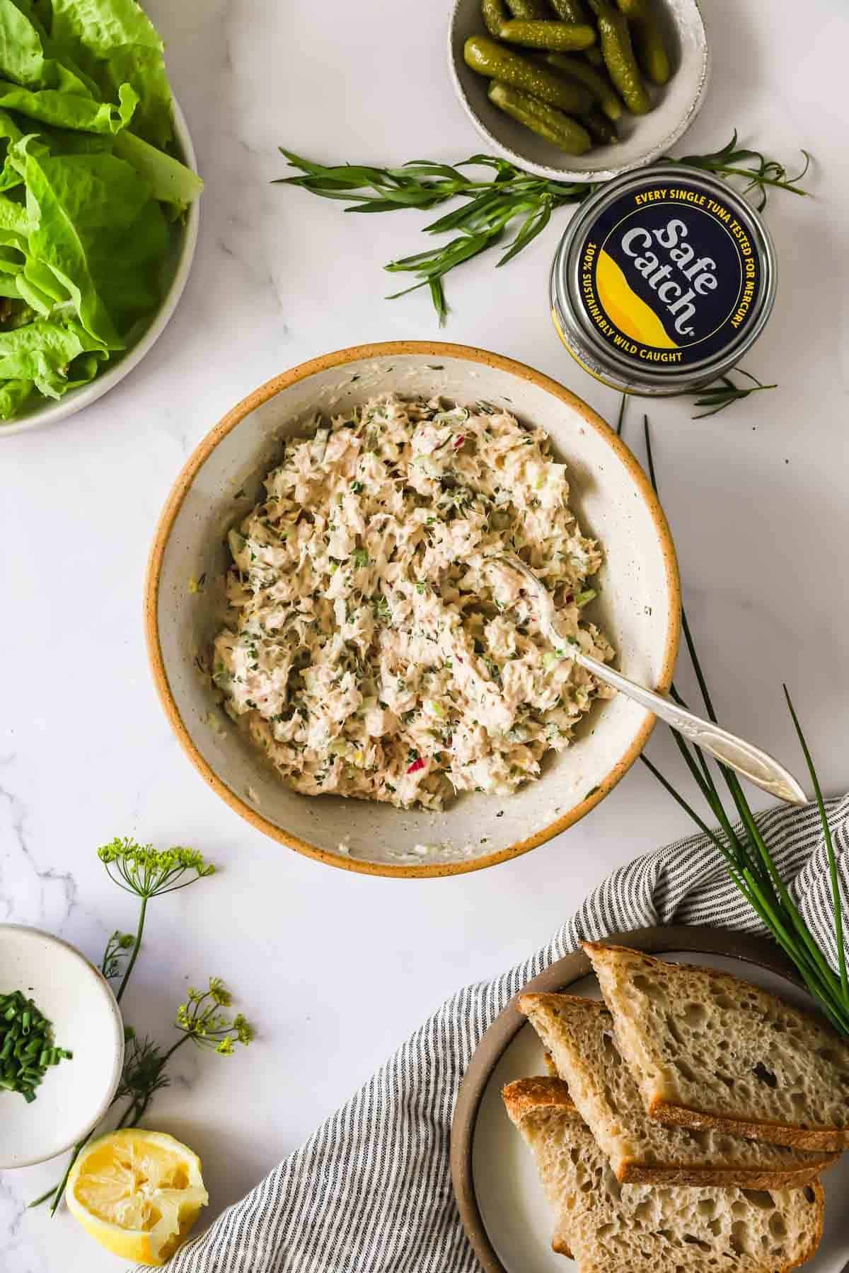 A bowl of tuna salad with a spoon, sliced bread, lettuce leaves, pickles, a can of tuna, and herbs on a white surface.