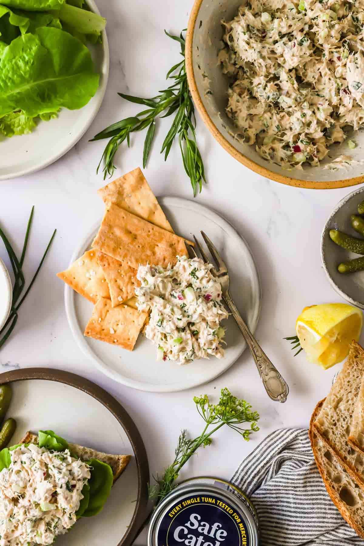 Plate with crackers topped with tuna salad beside herbs, lemon, lettuce, pickles, bread slices, and a bowl of tuna salad on a white surface.