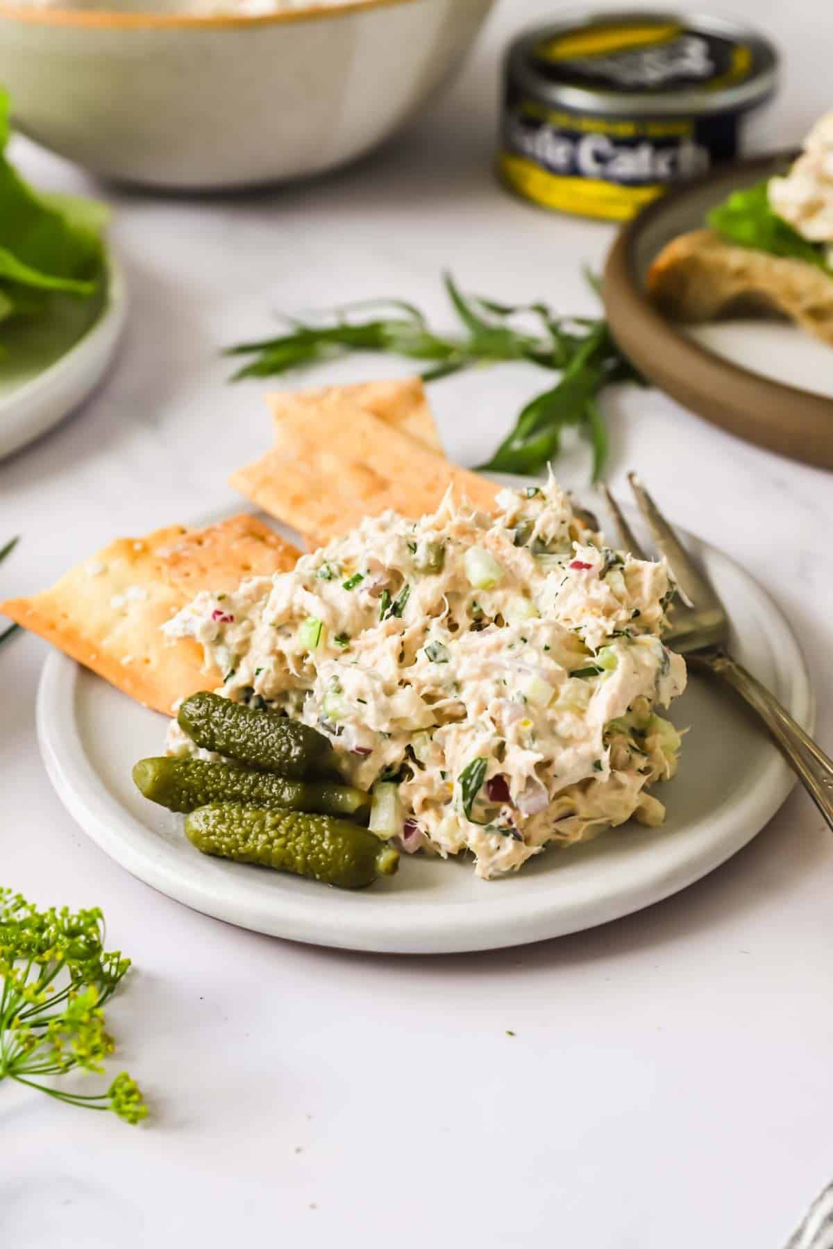 A plate with tuna salad, three small pickles, two crackers, and a fork. A can of tuna, bread, and greens are in the background.