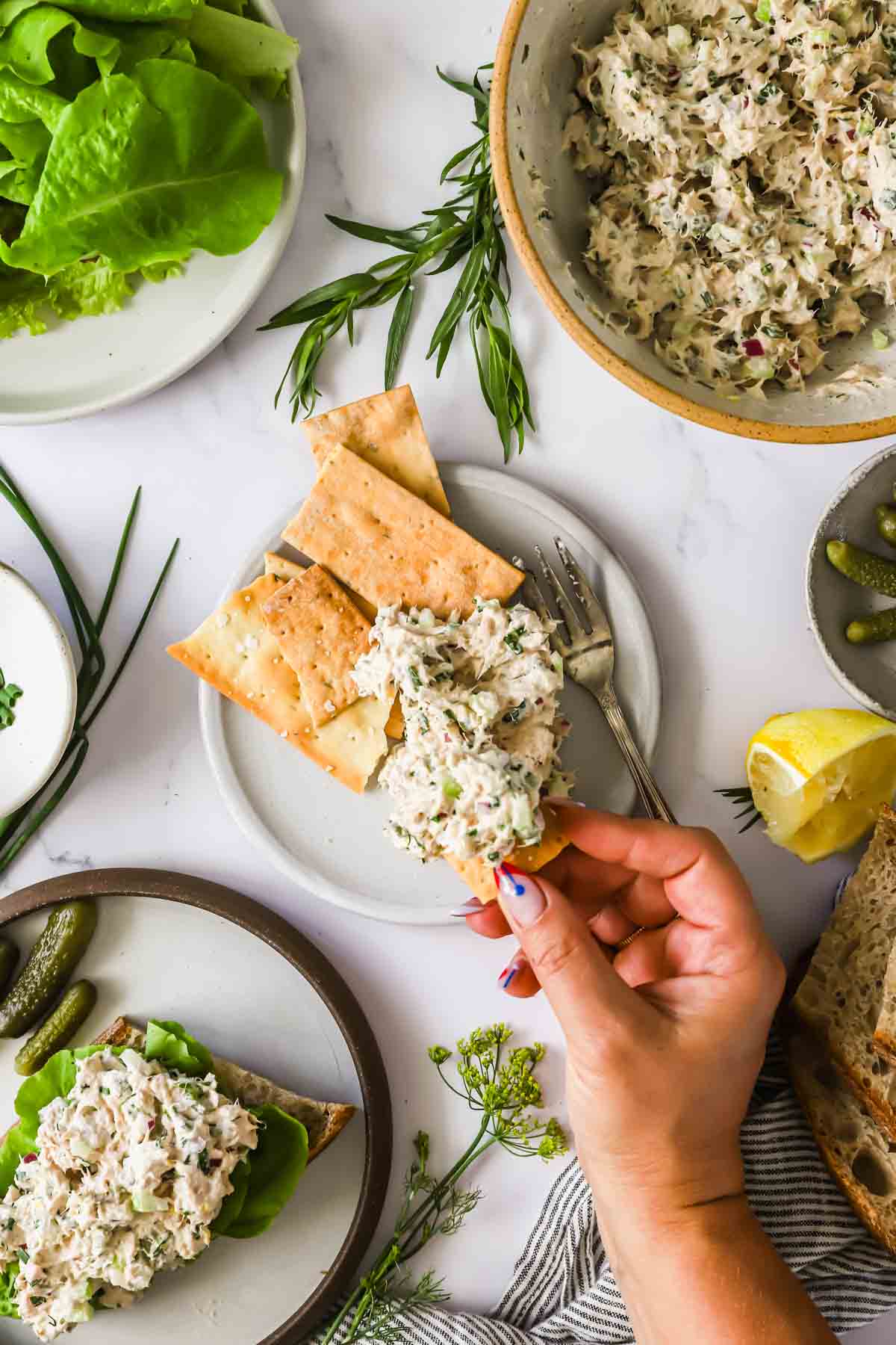 A hand spreads tuna salad with herbs on crackers, surrounded by bowls of salad, bread, fresh herbs, lettuce, lemon, and pickles on a white table.