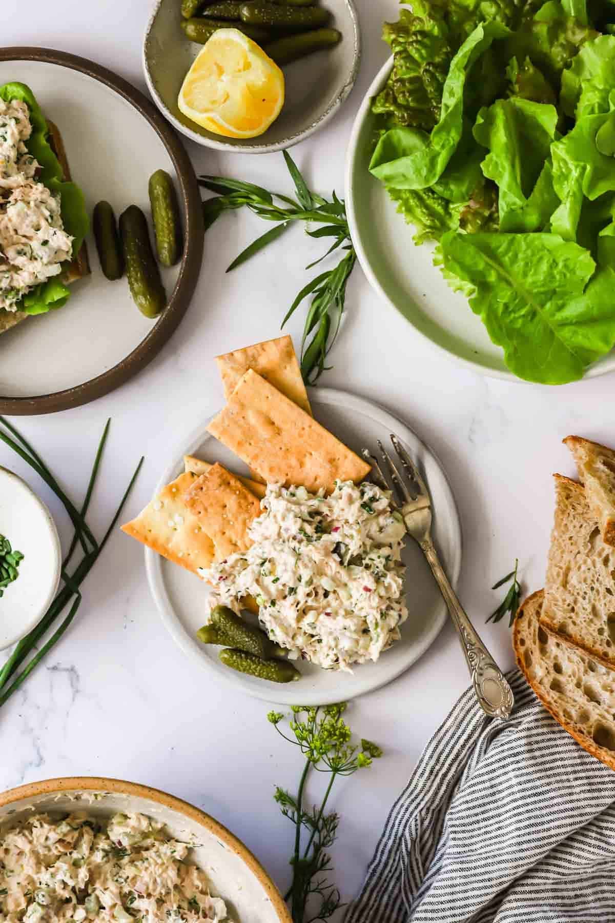 Plates with tuna salad, crackers, bread slices, pickles, lemon, and fresh greens arranged on a white surface.