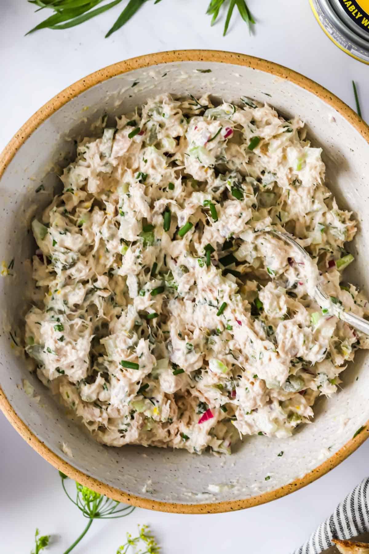 A bowl of tuna salad mixed with chopped celery, herbs, and mayonnaise, with a spoon resting inside the bowl.