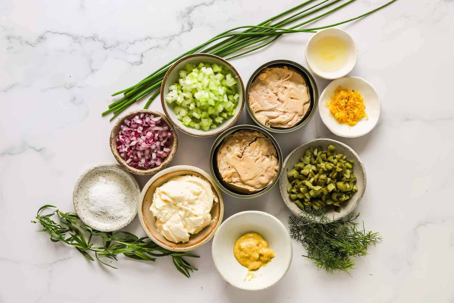 Bowls with ingredients including chopped pickles, celery, red onion, fresh herbs, mayonnaise, mustard, lemon juice, relish, and spices, arranged on a white marble surface.