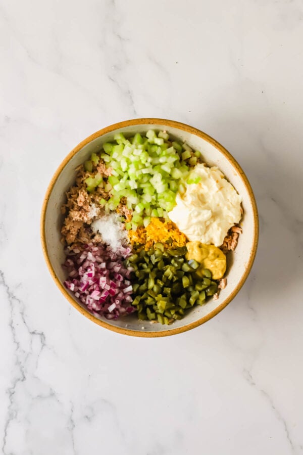A bowl containing chopped celery, red onion, pickles, mayonnaise, mustard, spices, and tuna, prepared for mixing on a marble surface.