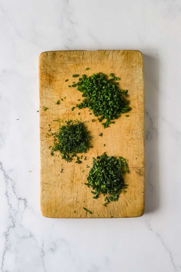 A wooden cutting board with three small piles of chopped green herbs on a white marble surface.