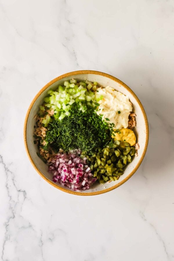 Bowl with chopped celery, red onion, pickles, herbs, mayonnaise, and mustard on a marble surface, ingredients unmixed.