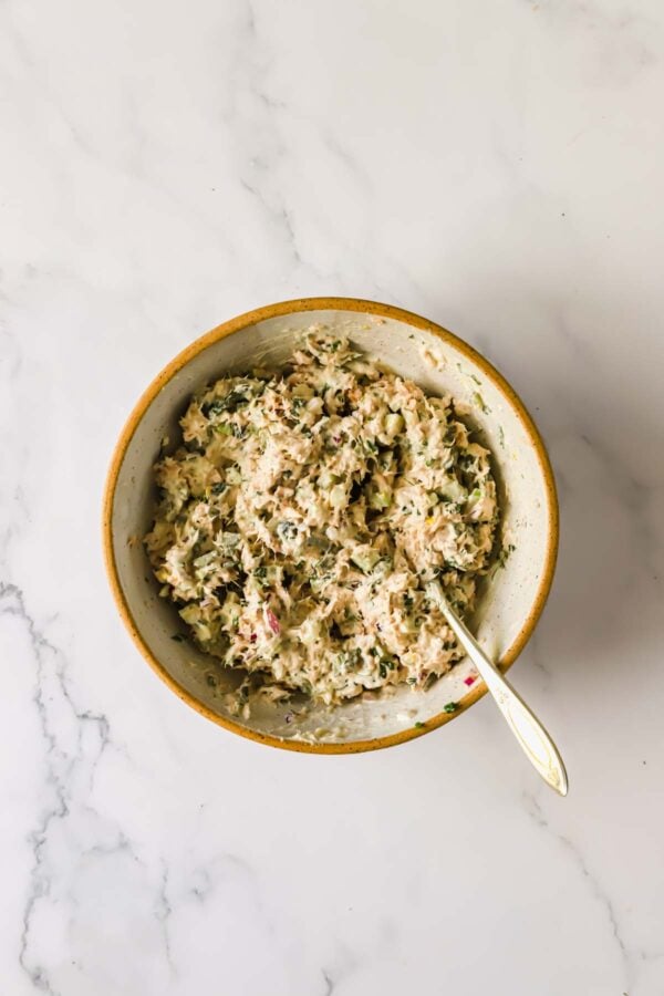 A ceramic bowl filled with a creamy tuna salad mixture, featuring herbs and chopped vegetables, with a spoon resting inside.