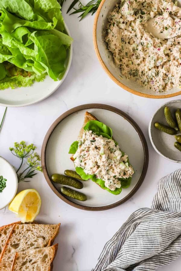 Open-faced chicken salad sandwich on a plate with lettuce and pickles, surrounded by a bowl of chicken salad, fresh lettuce, sliced bread, lemon, and a striped napkin.