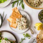 A plate with crackers and a serving of tuna salad, surrounded by herbs, a lemon wedge, pickles, and bread on a white surface.