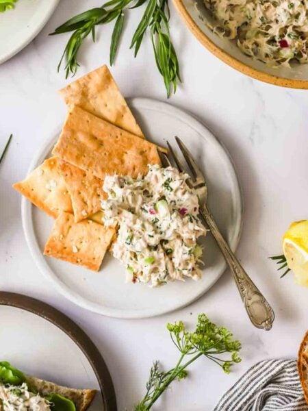 A plate with crackers and a serving of tuna salad, surrounded by herbs, a lemon wedge, pickles, and bread on a white surface.