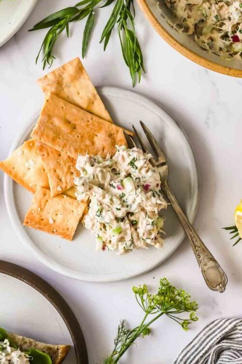 A plate with crackers and a serving of tuna salad, surrounded by herbs, a lemon wedge, pickles, and bread on a white surface.