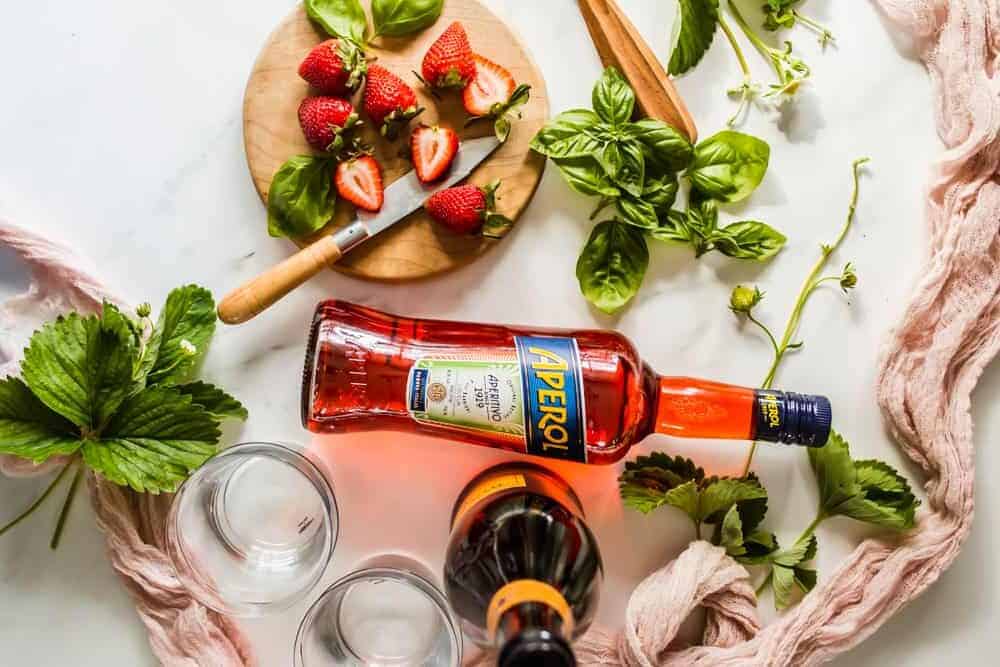 bottles of Aperol and prosecco next to basil leaves and a wood board with strawberries