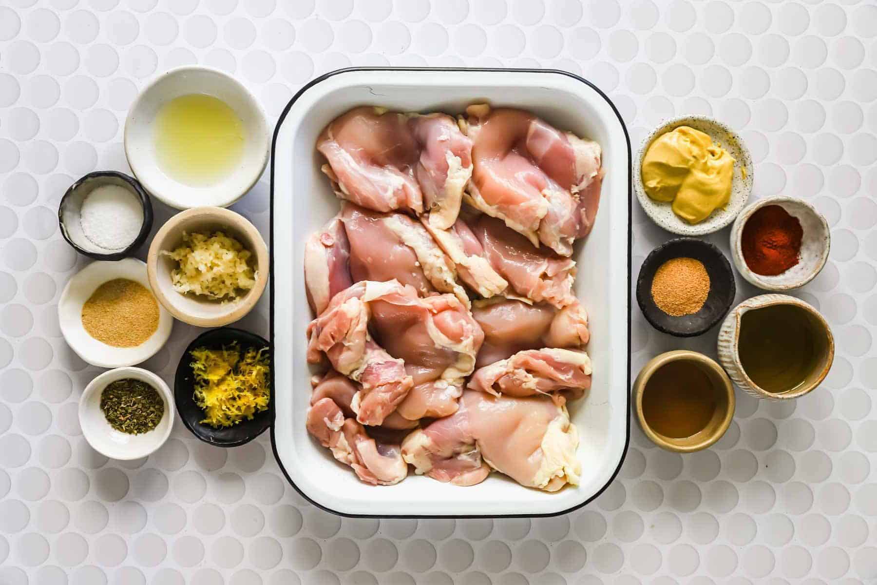 Raw chicken thighs in a white tray surrounded by small bowls of spices, mustard, oil, minced garlic, lemon zest, and seasonings on a white textured surface.