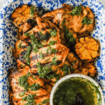 Grilled chicken thighs topped with green herb sauce, served with charred lemon halves in a blue speckled dish, alongside a small bowl of extra sauce and a spoon.