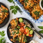 Grilled chicken thighs with herbs served on plates with a cucumber and tomato salad, grilled lemon, and fresh parsley on a white table.