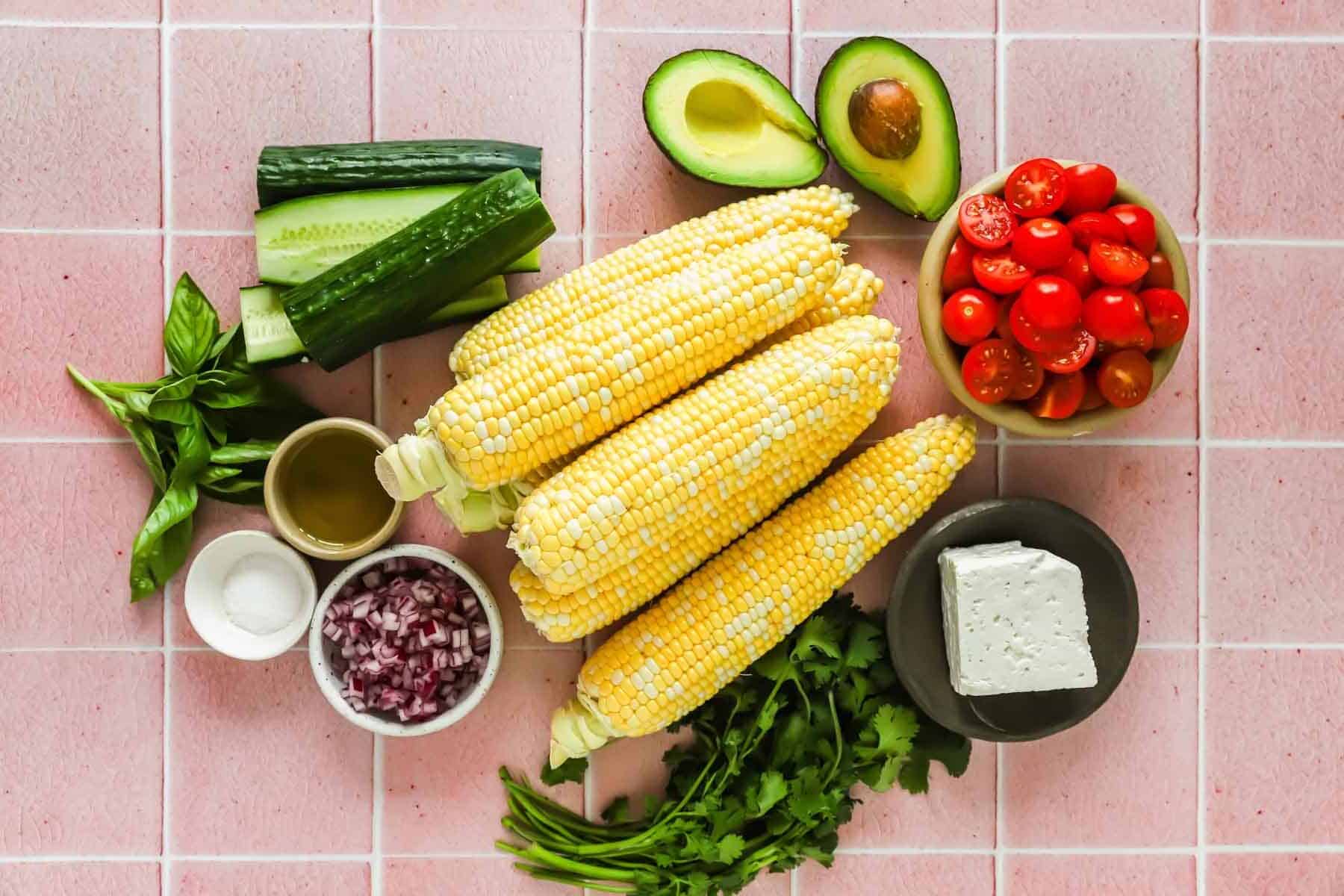 Fresh ingredients on a pink tile surface: corn, avocado, grape tomatoes, cucumber, red onion, cilantro, basil, feta cheese, olive oil, and salt.