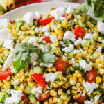 A plate of grilled corn salad with tomatoes, cucumber, cilantro, and crumbled cheese, placed on a pink surface with cilantro and grilled corn in the background.