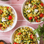 Three bowls of grilled corn salad with cherry tomatoes, cucumber, cheese, and herbs on a pink tile surface, surrounded by lime wedges, spices, and a bowl of crumbled cheese.