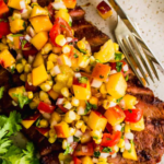 Grilled flank steak topped with a colorful peach and corn salsa, served on a white plate with a fork and parsley garnish.