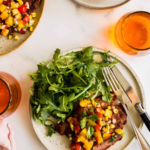 Plate with grilled flank steak topped with mango salsa and arugula, served with a fork and glass of orange drink on a white surface.