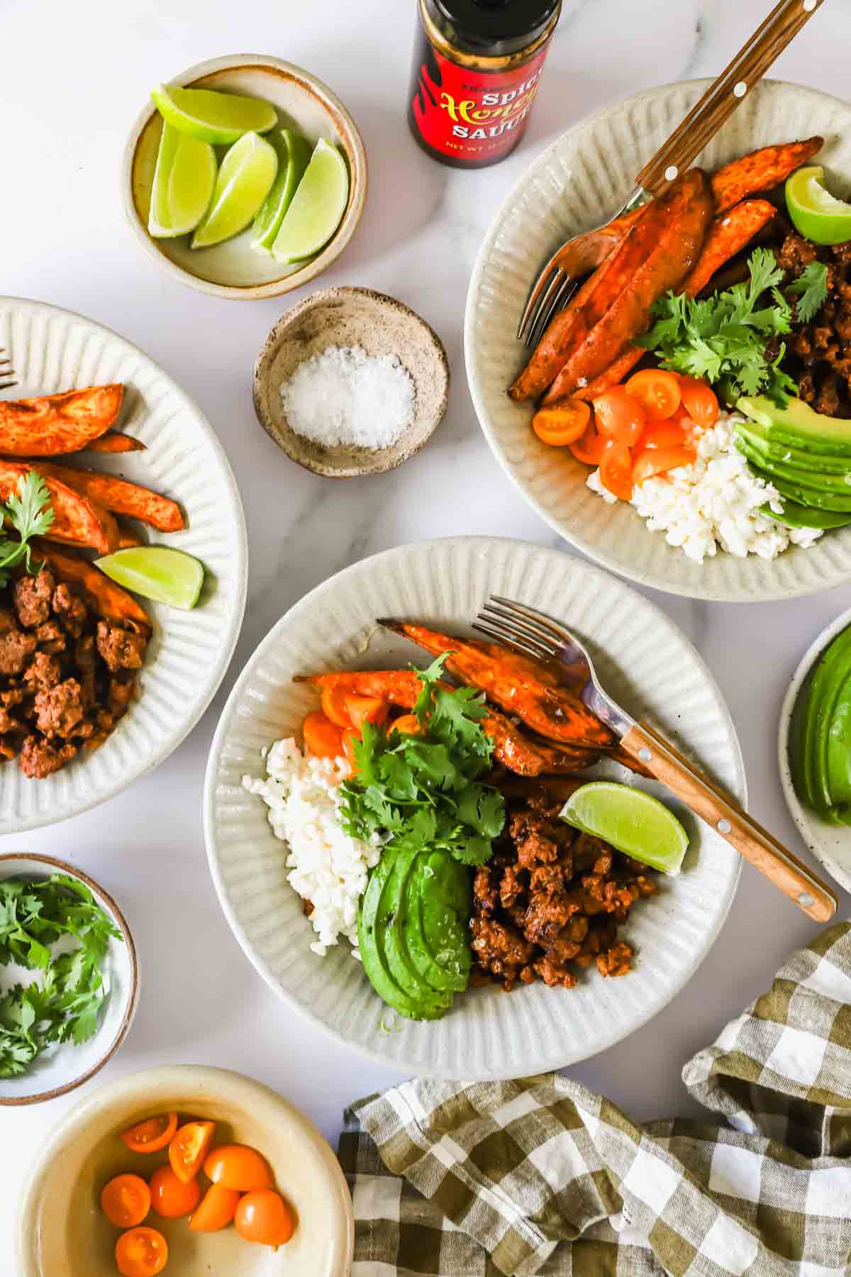 Three plates with rice, ground meat, roasted sweet potatoes, avocado, cherry tomatoes, and lime wedges. Small bowls of limes, salt, cilantro, tomatoes, and a bottle of Sriracha are nearby.