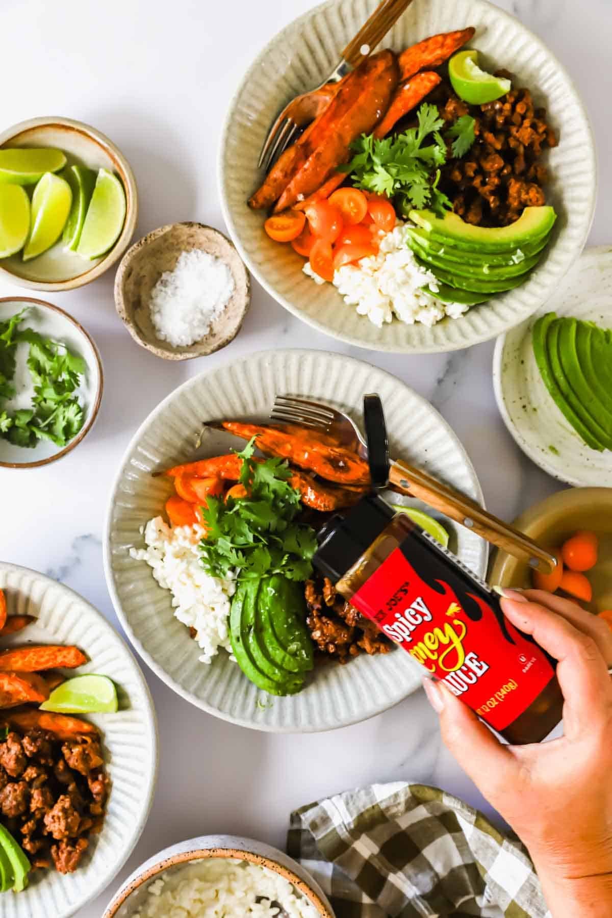 Three bowls with rice, sliced avocado, sweet potatoes, cilantro, and ground meat; one hand holds a bottle of spicy sauce. Sliced limes, cilantro, and salt in small bowls are nearby.