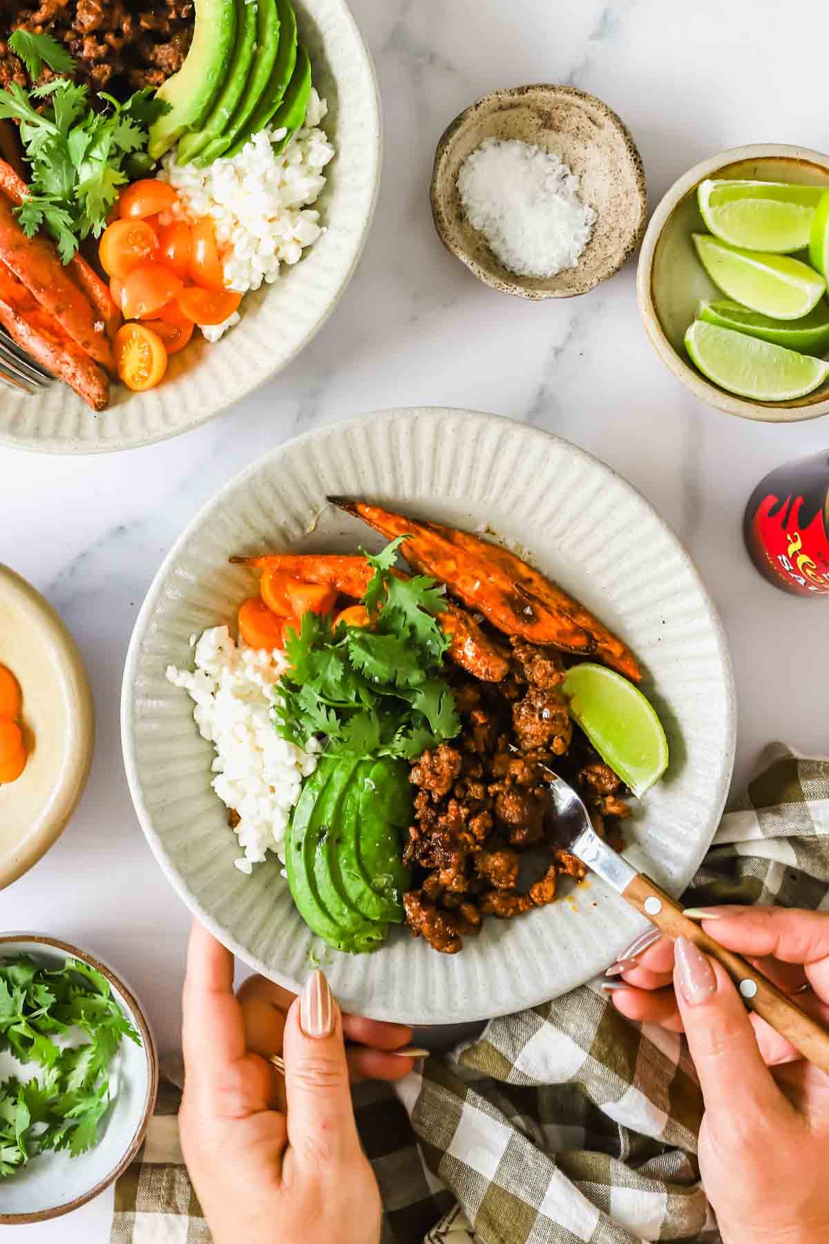 A hand holds a plate with rice, seasoned ground meat, sliced avocado, carrots, cilantro, a lime wedge, and roasted carrots. Bowls of cilantro, lime, and salt are nearby on a marble surface.