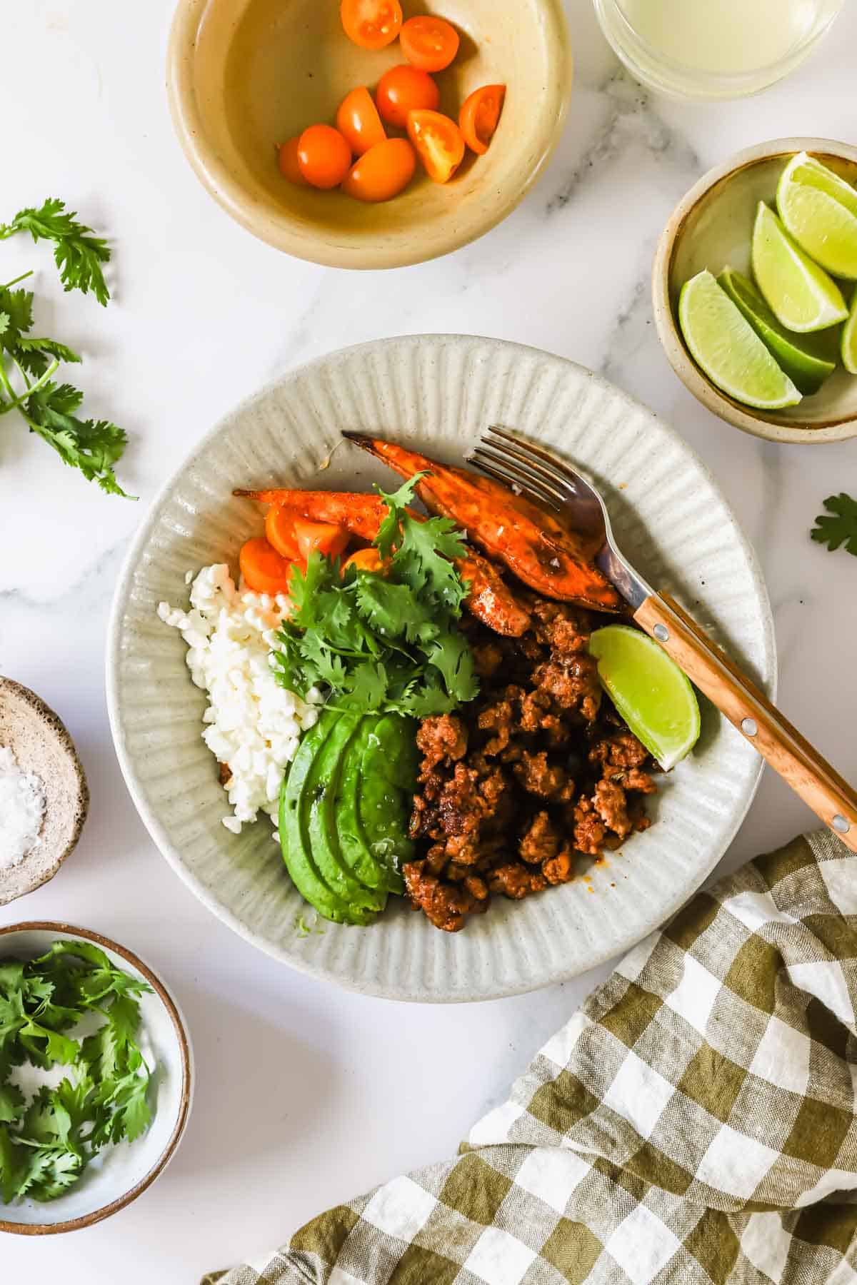 A bowl with rice, sliced avocado, cooked ground meat, carrot, fresh cilantro, and a lime wedge, with small bowls of tomatoes, lime wedges, and cilantro nearby on a white surface.